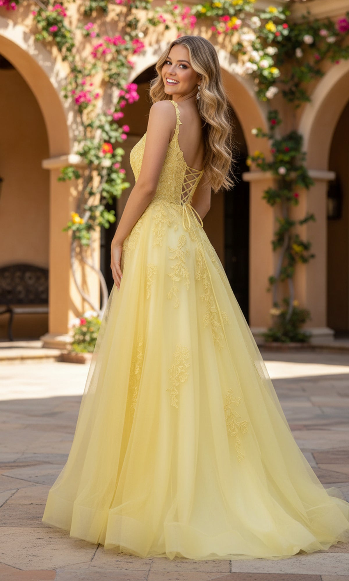 A woman in the Long Prom Dress: PromGirl Exclusive 250261, a yellow floor-length A-line gown, stands outdoors on a tiled patio with arches and flowering vines in the background.